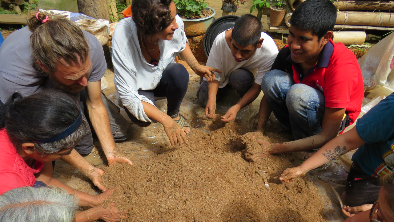 Six people are crouching down in a circle looking at fertiliser they have made for the coffee plants. They are outside. Behind them are organic agricultural materials. The woman in the centre of the photo is talking animatedly about the fertiliser to the two men to her right. 