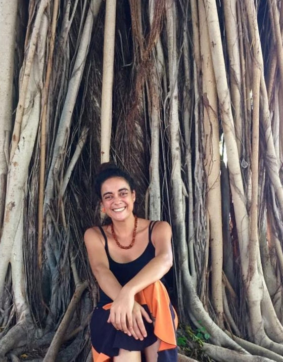 A woman couches at the base of intertwined trees. She has her black hair pulled up, a bead necklace, black singlet and orange skirt.