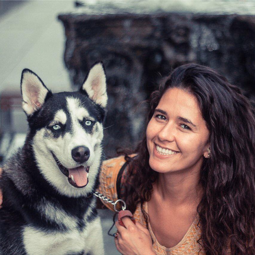 A woman with long dark hair smiles, and holds the lead of a husky dog