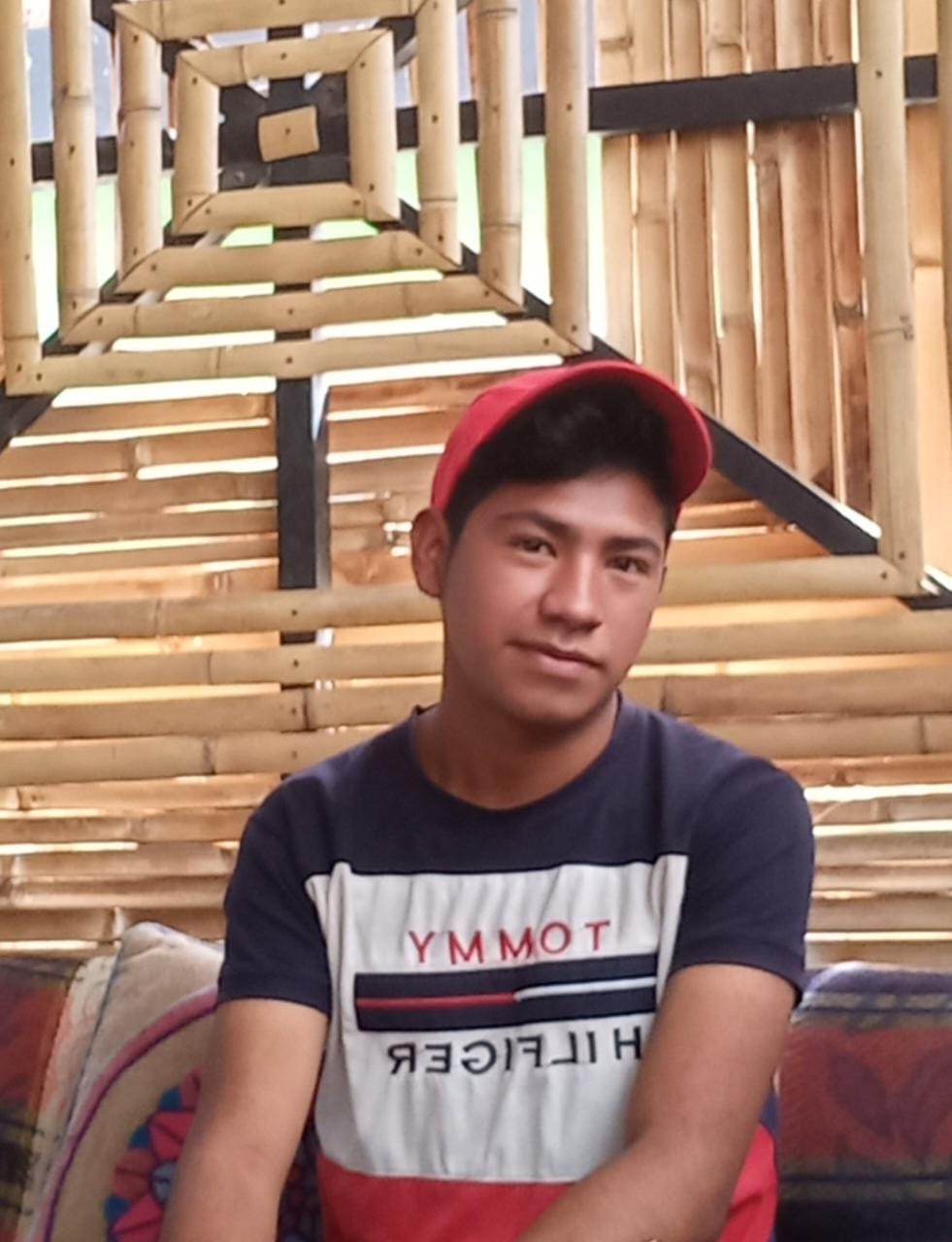 A young man sits in front of a bamboo wall. He wears a cap and teeshirt