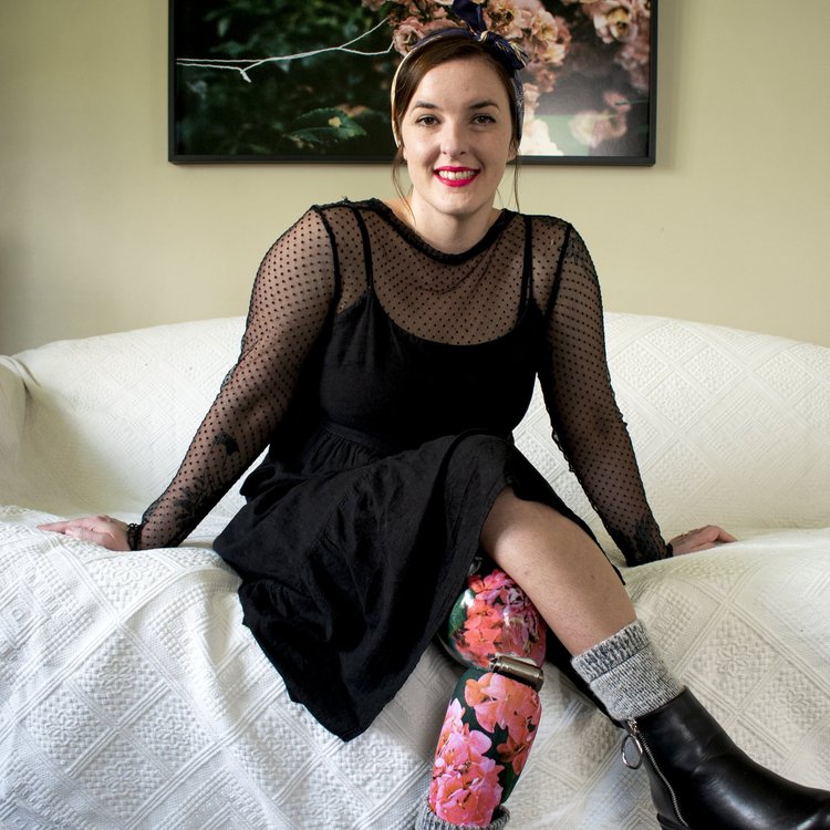A woman sits on a white couch. She wears a black dress and a floral prosthetic leg.