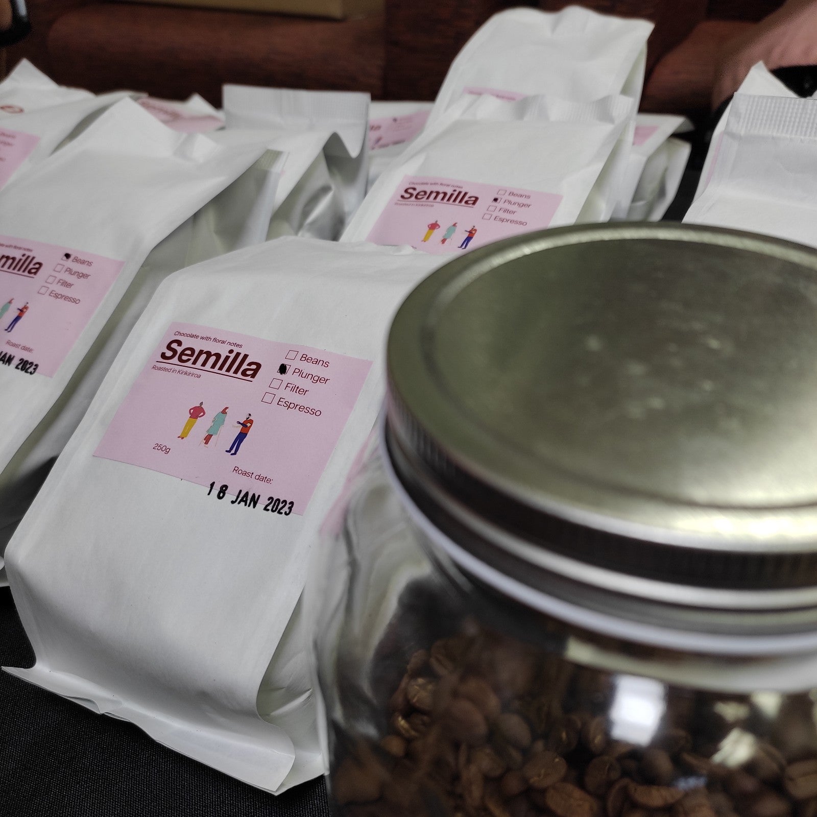 Close up of two white bags of Semilla coffee. The pink Semilla label is clear on the second bag. It says ‘Chocolate with floral notes, Semilla, Roasted in Kirikiriroa. 250g. Beans, plunger, filter, espresso. Roast date: 18 Jan 2023. The box next to plunger is marked. There is a grey lid next to the two bags on the right-hand side of the picture. 